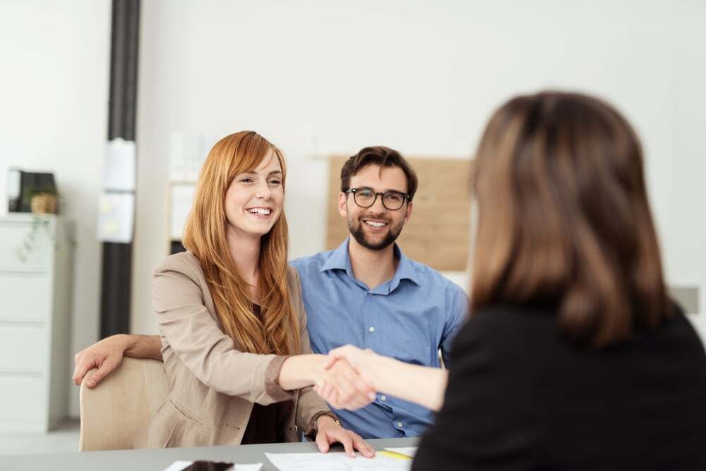 Happy young couple meeting with a broker in her office leaning over the desk to shake hands, view from behind the female agent-Oct-18-2024-05-43-21-0988-AM-1