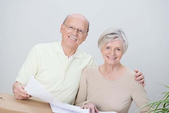 Smiling affectionate senior couple standing with the husbands arm around his wife looking at documents resting on a brown cardboard carton as they plan their move to a new home-4-1